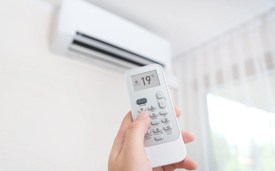 Mano sosteniendo un control remoto ajustando la temperatura de un aire acondicionado tipo minisplit montado en la pared en un ambiente moderno y luminoso.